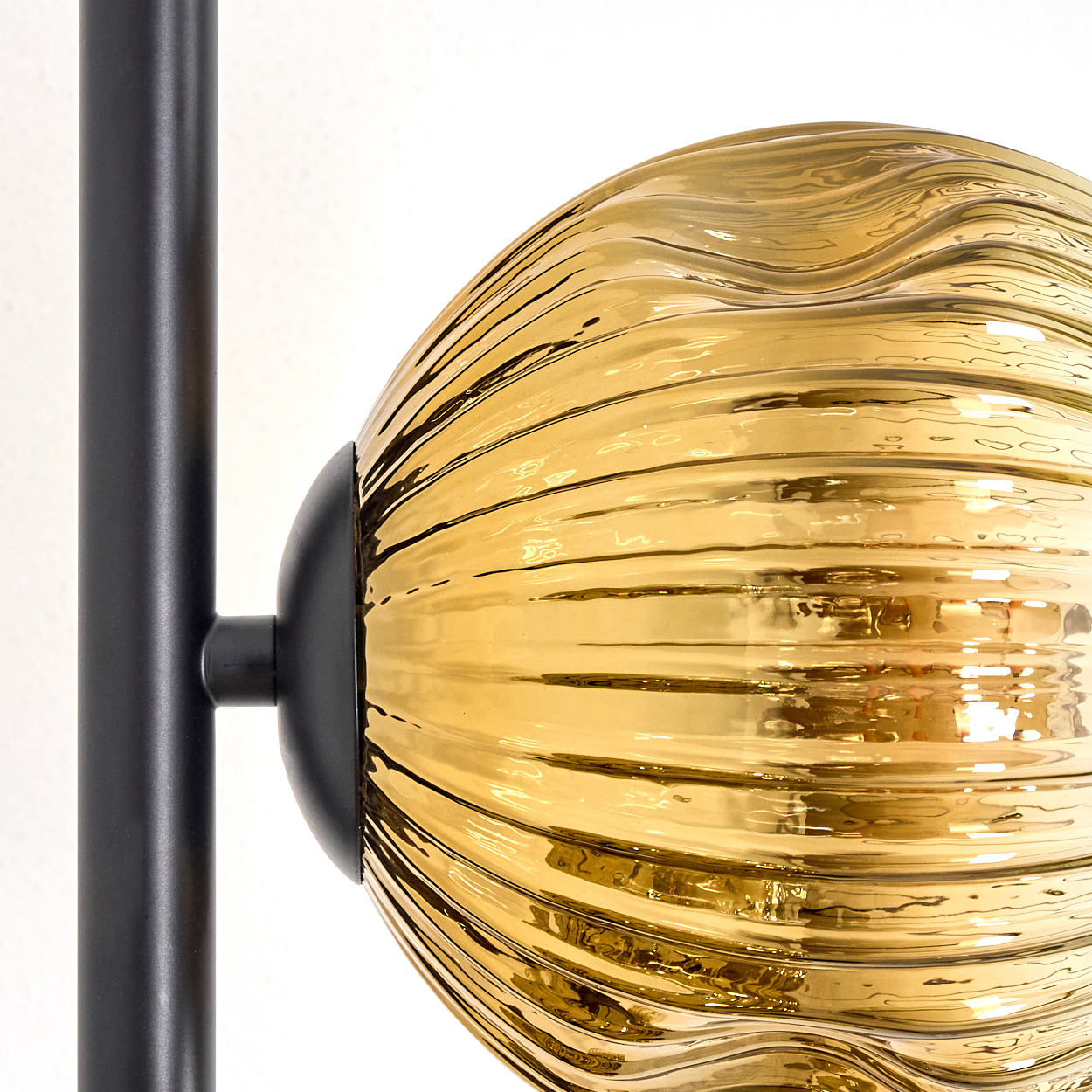Nahaufnahme einer Lampe. Ein schwarzer Metallrahmen trägt eine goldene, strukturierte Glaskugel. Der Hintergrund ist eine weiße Wand.