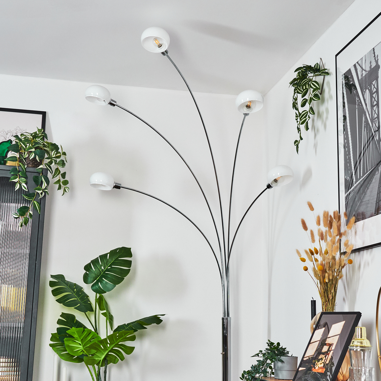 Eine moderne Stehlampe mit fünf weißen Kugellampen. Pflanzen und ein gerahmtes Bild schmücken den Raum mit weißen Wänden.