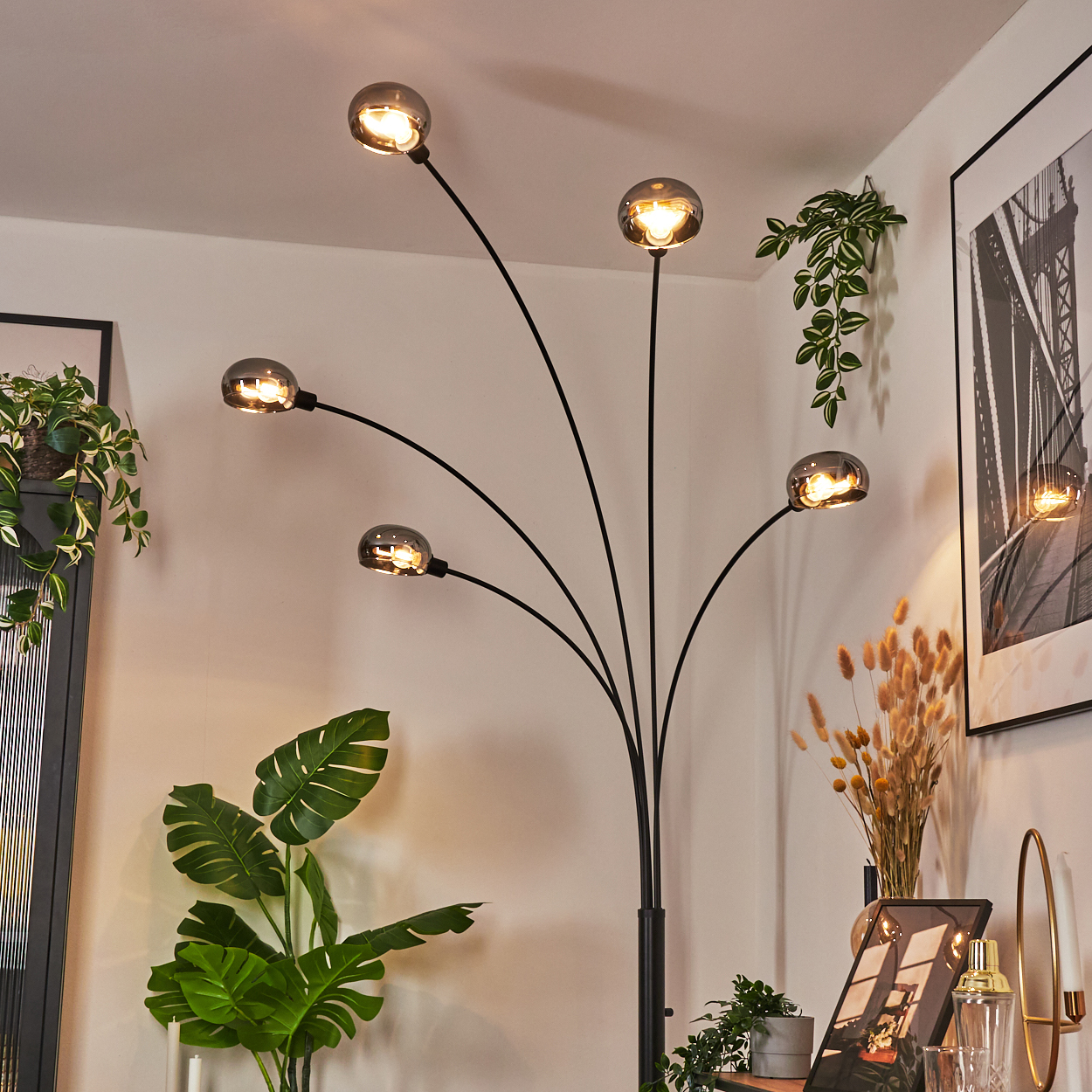Eine moderne Stehlampe mit fünf Lichtern. Pflanzen und Kunstwerke umgeben die Lampe in einem gut beleuchteten Innenraum. Die Farbpalette ist neutral.