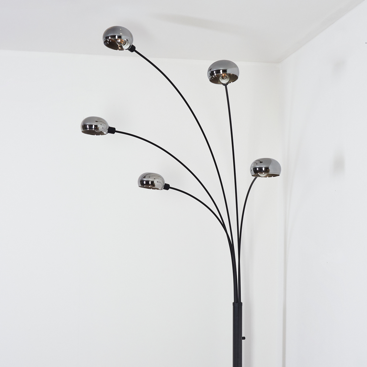 Eine moderne Stehlampe mit fünf verchromten Kuppellampen und schwarzen Stielen vor weißen Wänden in einer Ecke.