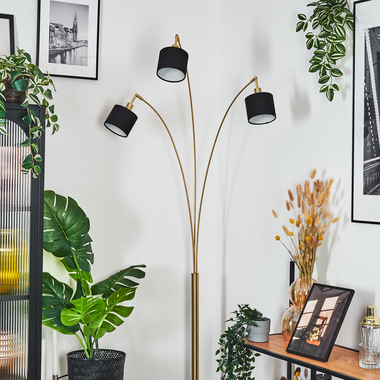 Ein Raum mit einer goldenen Stehlampe mit schwarzen Schirmen, Pflanzen, Bildern und einem Regal mit Dekoration. Die Wände sind weiß.