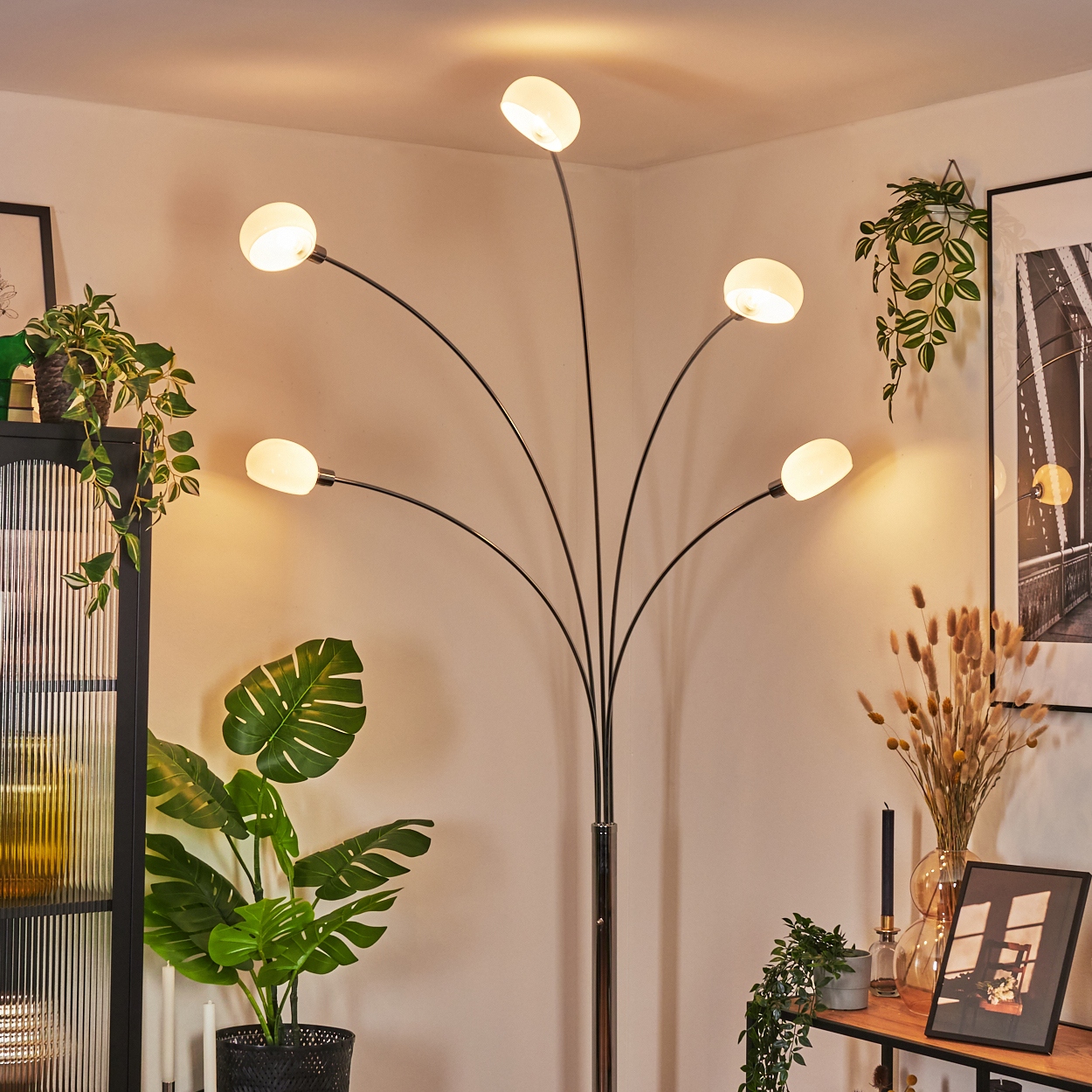 Eine moderne Stehlampe mit fünf weißen Kugelleuchten. Pflanzen und ein gerahmtes Bild schmücken den Raum, links ein schwarzer Schrank.