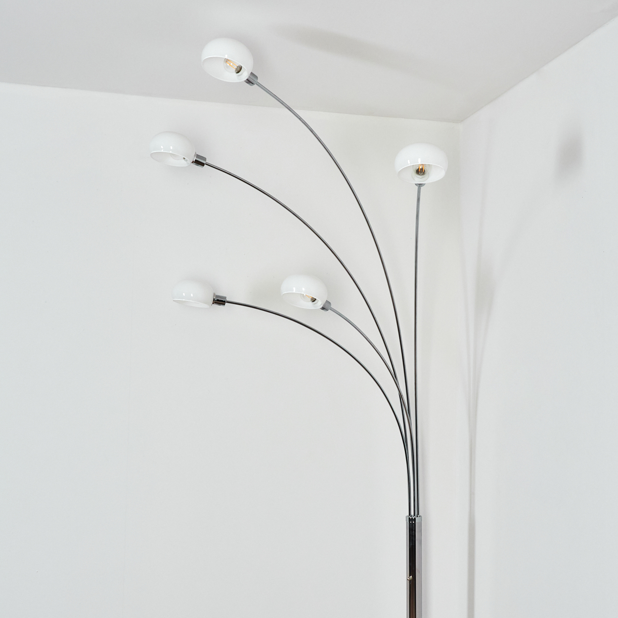 Eine moderne Stehlampe mit fünf weißen Kugelleuchten an gebogenen Armen. Sie steht in einer weißen Ecke und wirft einen Schatten.