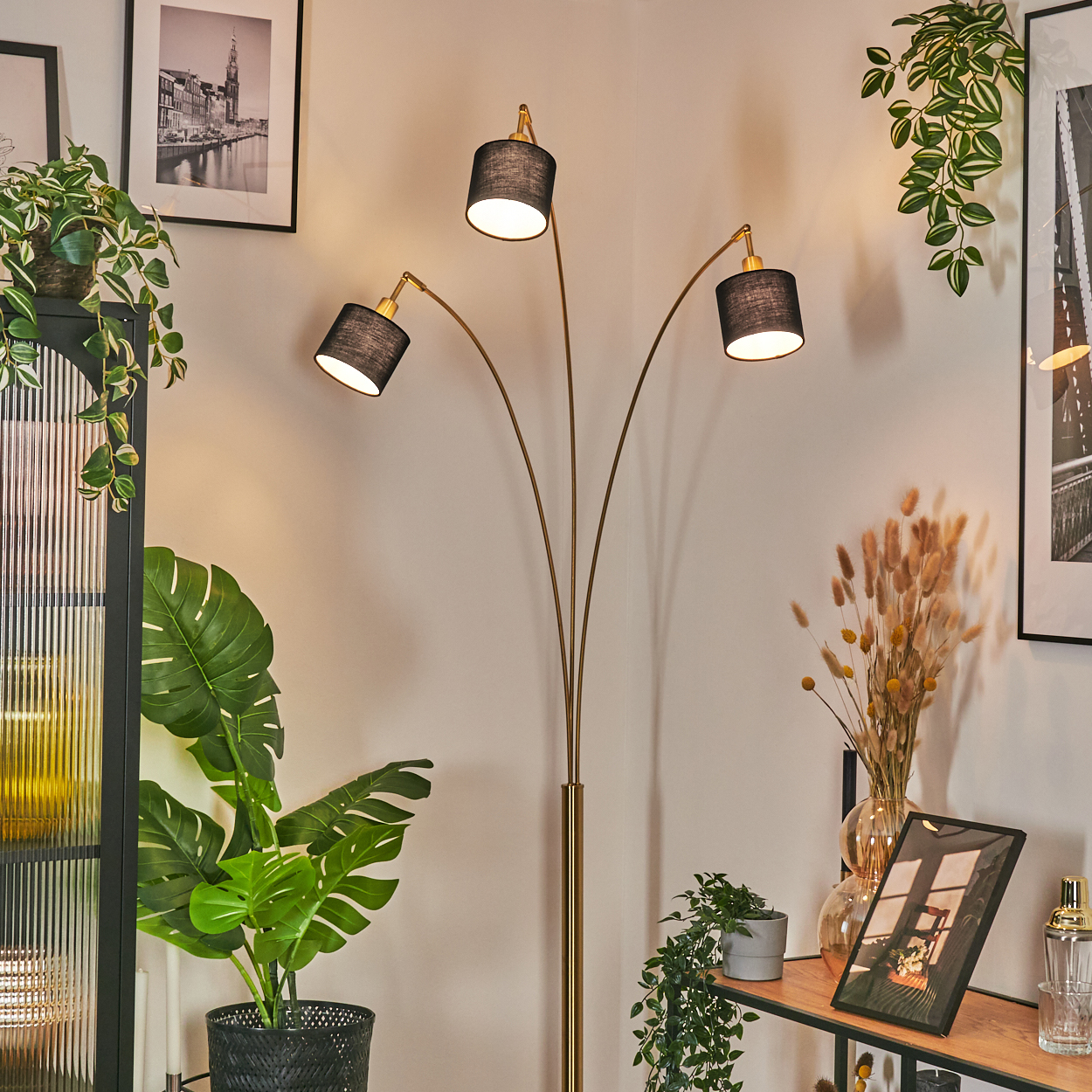 Eine goldfarbene Stehlampe mit drei schwarzen Schirmen. Pflanzen in Töpfen und gerahmte Bilder schmücken den Raum. Die Wände sind hellbeige.