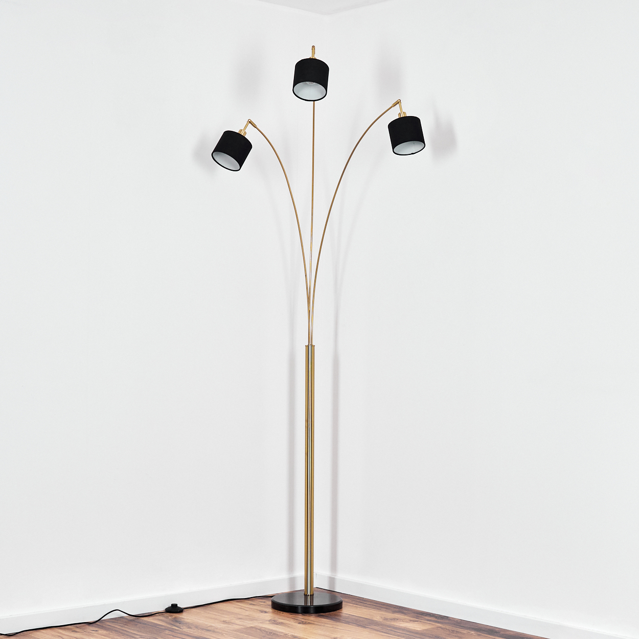 Eine goldene Stehlampe mit drei schwarzen Lampenschirmen steht in einer weißen Ecke. Der Boden ist aus Holz.