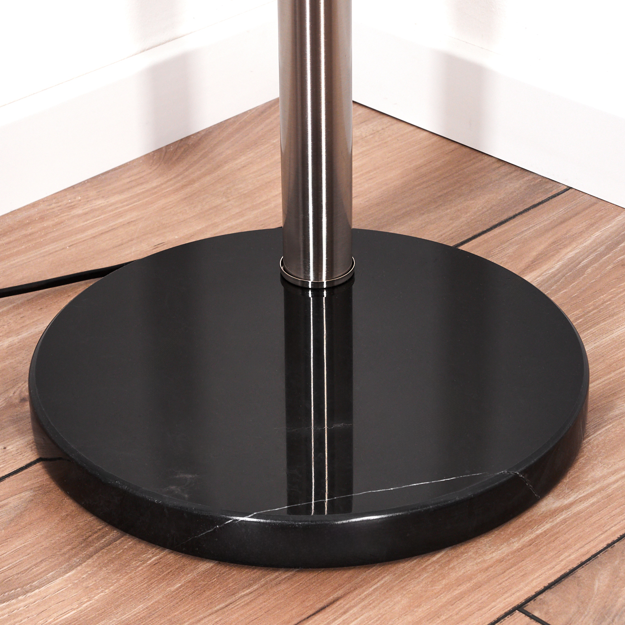 Nahaufnahme eines Lampenfußes. Eine schwarze, kreisförmige Basis trägt eine silberne Stange. Die Basis steht auf Holzböden, im Hintergrund eine weiße Wand.