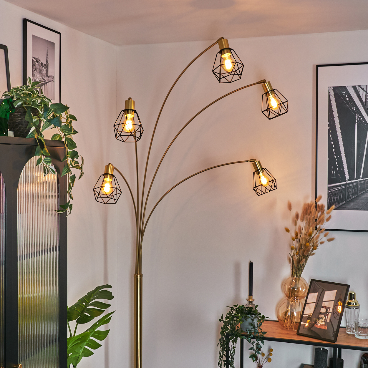 Eine moderne Stehlampe mit fünf Lichtern, Pflanzen und gerahmten Bildern. Die Lampe ist goldfarben und die Schirme sind schwarzer Draht.