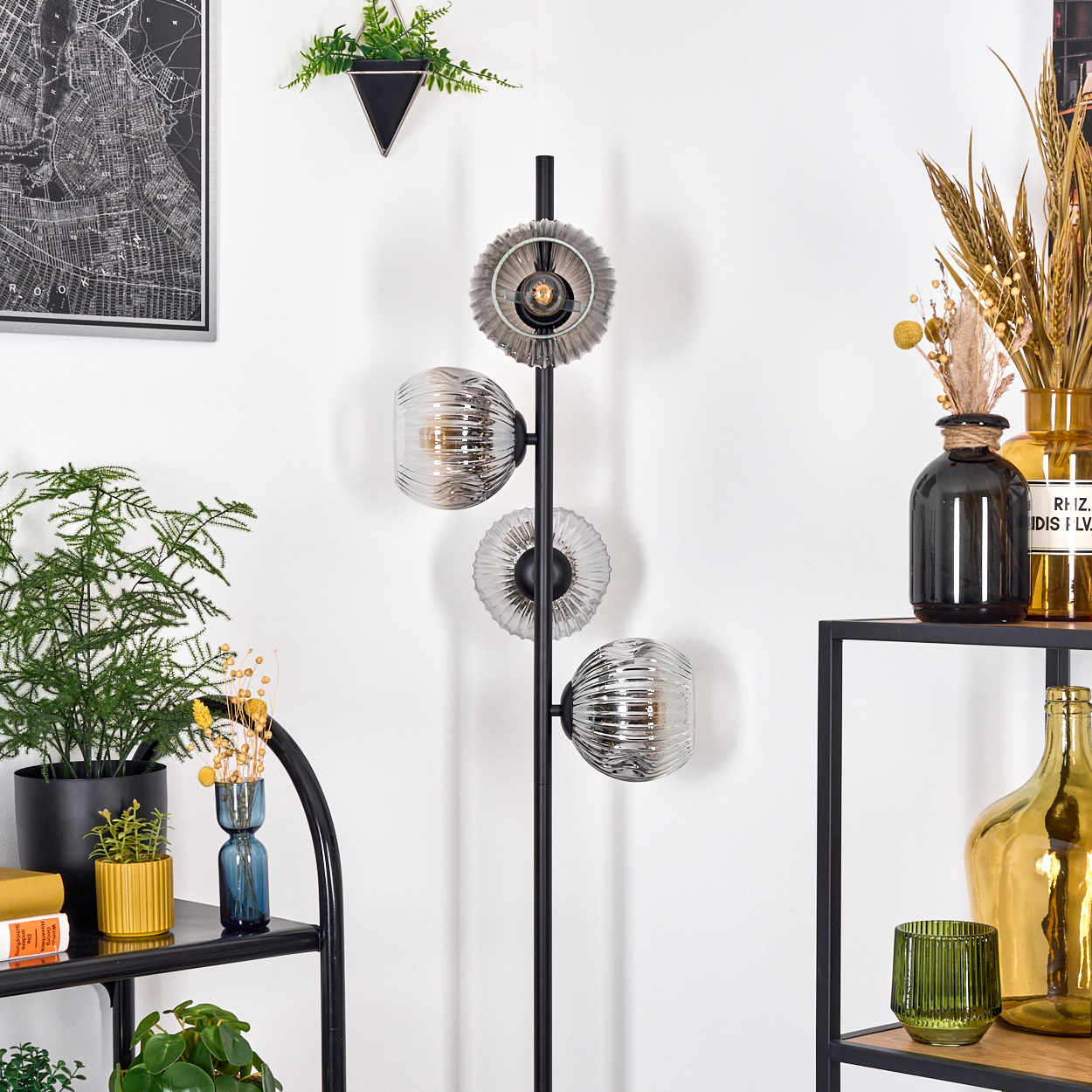 Eine moderne Stehlampe mit drei Glasschirmen. Regale mit Pflanzen, Vasen und einer Karte hängen an der weißen Wand.