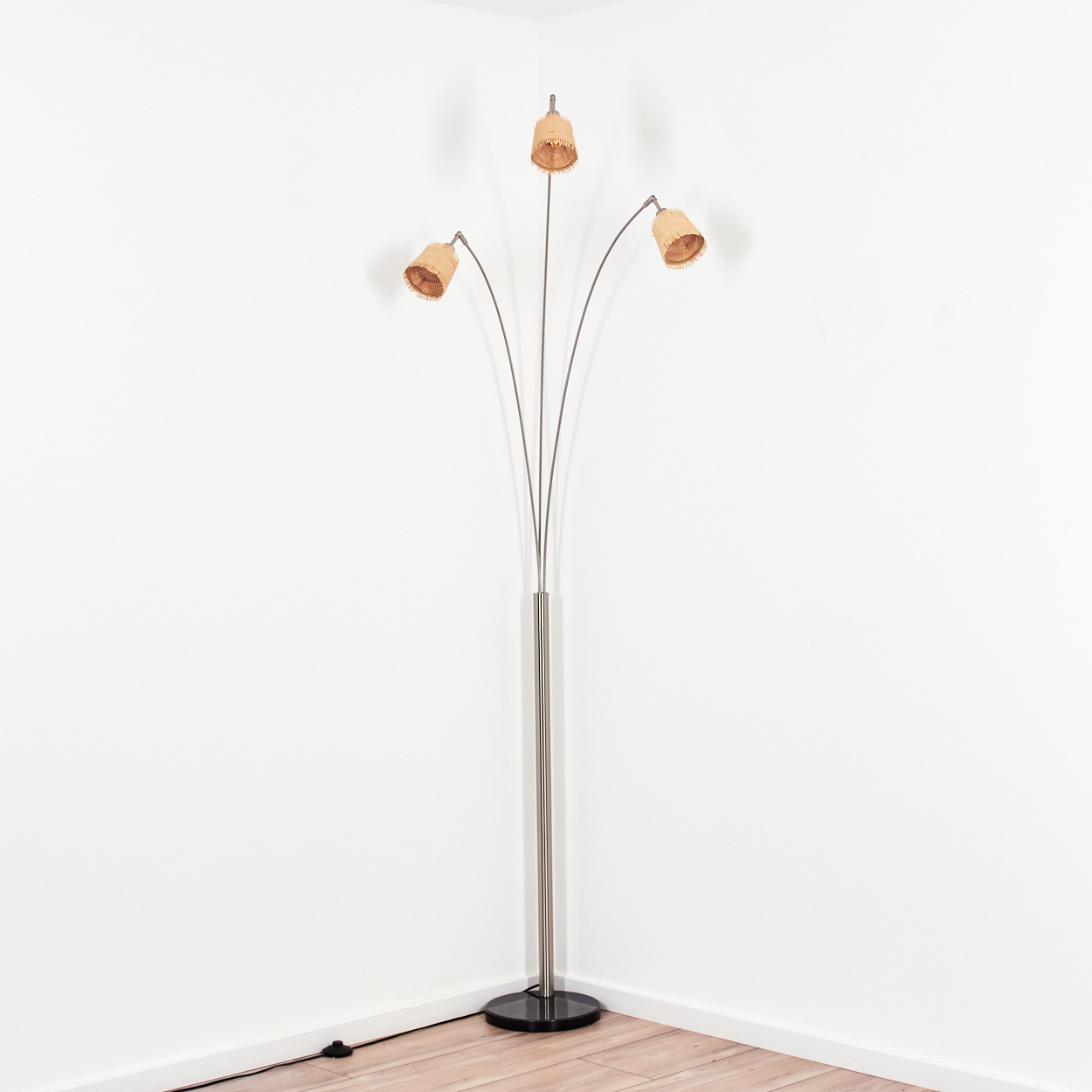 Eine Stehlampe mit Metallstiel und drei Lampenschirmen. Die Schirme sind beige und der Sockel ist schwarz. Sie steht in einer Ecke.