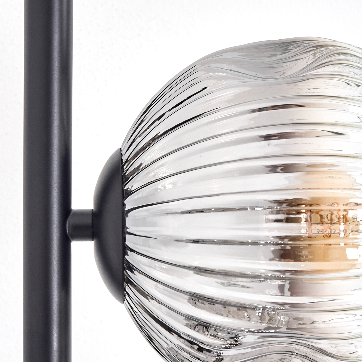 Nahaufnahme einer modernen Lampe. Eine klare, geriffelte Glaskugel ist an einer schwarzen Metallstange befestigt, vor weißem Hintergrund.
