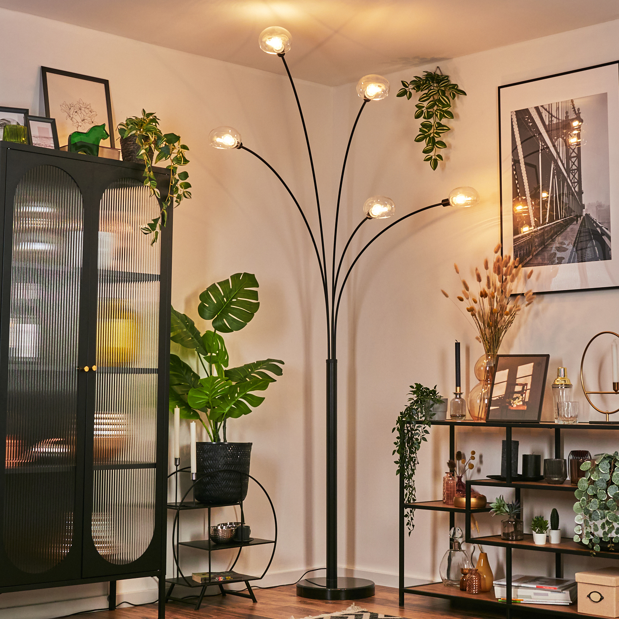 Ecke eines Wohnzimmers mit schwarzer Stehlampe, Pflanzen und Regalen mit Dekorationen. Ein gerahmtes Brückenfoto hängt an der Wand.