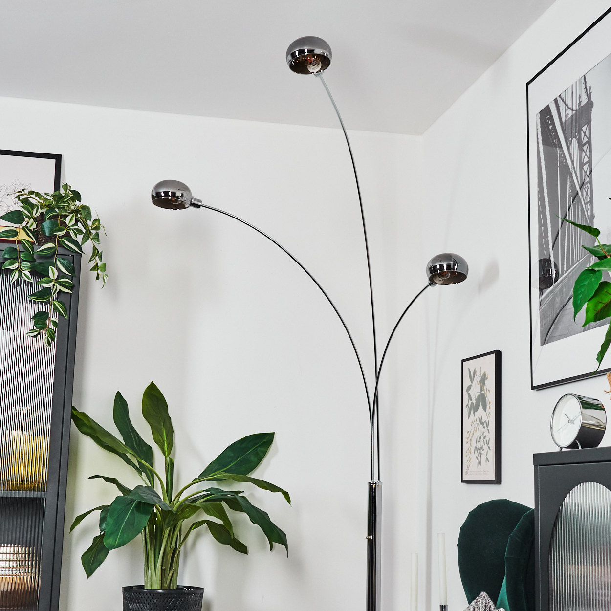 Eine moderne Stehlampe mit drei verchromten Kugelleuchten. Pflanzen, ein schwarzer Schrank und gerahmte Kunstwerke im Hintergrund.
