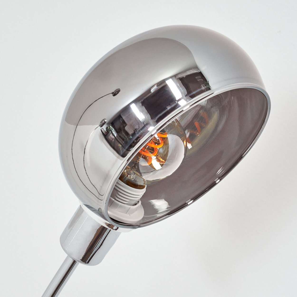 Eine Chromlampe mit Glühbirne. Der Glühfaden ist sichtbar. Die Lampe steht vor weißem Hintergrund und reflektiert das Licht.