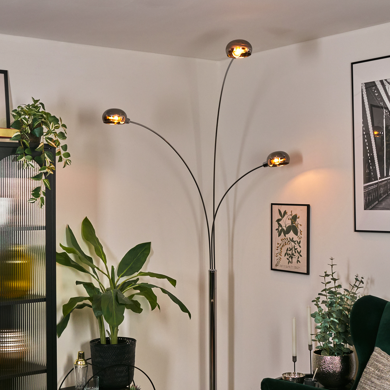 Eine moderne Stehlampe mit drei Köpfen beleuchtet einen Raum mit Pflanzen, einem Bild und einem grünen Sessel.
