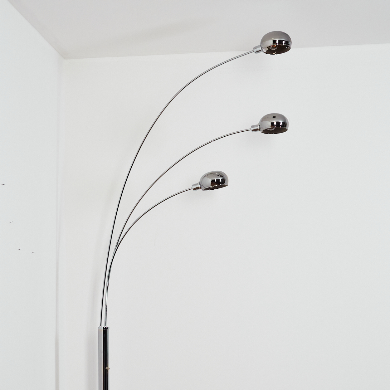 Eine verchromte Stehlampe mit drei gebogenen Armen, die jeweils ein rundes Licht halten. Die Lampe steht vor einer weißen Wand in einer Ecke.