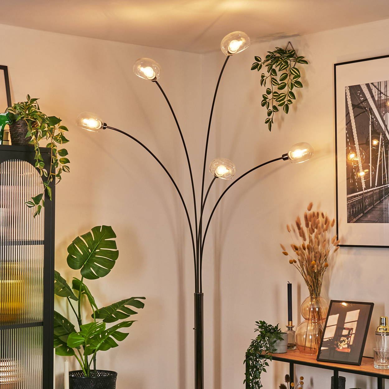 Ein modernes Wohnzimmer mit einer schwarzen Stehlampe mit fünf Glühbirnen. Pflanzen und gerahmte Bilder schmücken den Raum. Die Wände sind hell.