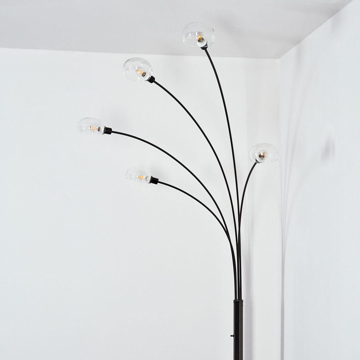 Eine moderne Stehlampe mit fünf gebogenen schwarzen Stielen, die jeweils eine klare Glühbirne halten. Die Lampe steht in einer weißen Ecke.