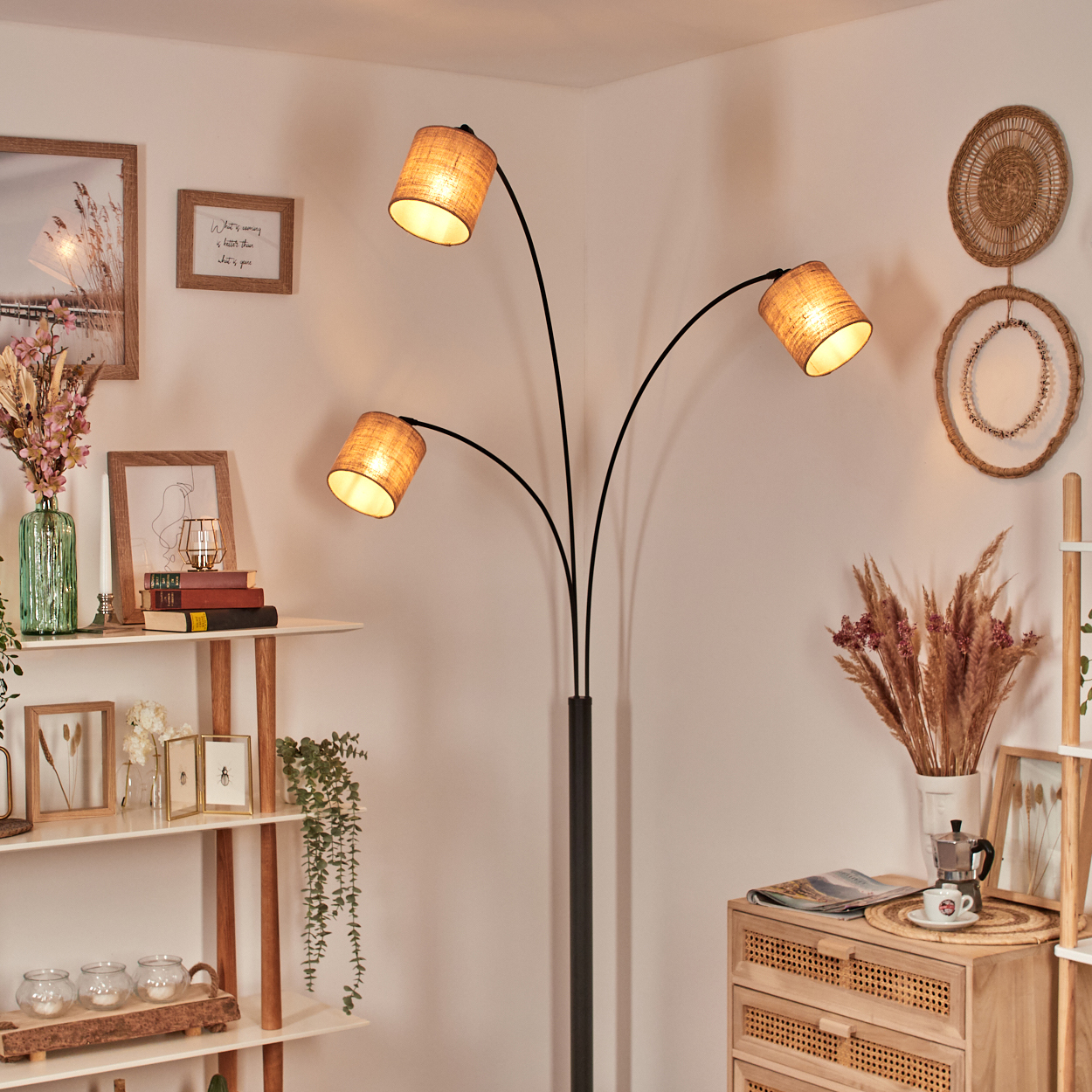 Ein Raum mit Stehlampe, Regalen mit Deko und einer Holzkommode. Die Lampe hat drei Schirme. Der Raum ist warm beleuchtet.