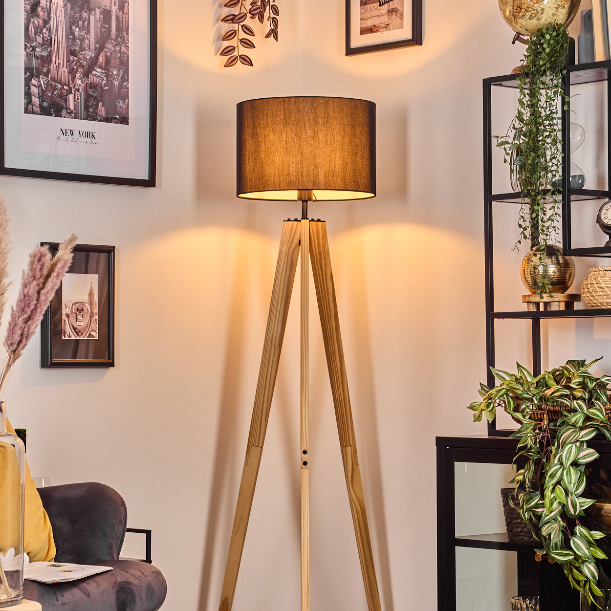Ein Raum mit einer hölzernen Stativlampe, gerahmter Kunst und einem schwarzen Bücherregal mit Pflanzen und Dekoration.