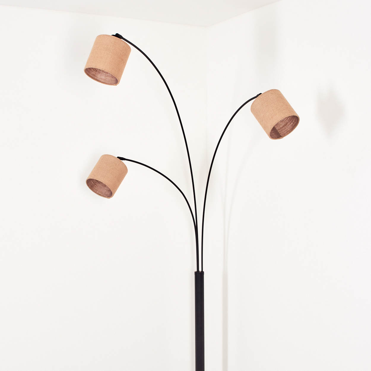 Eine moderne Stehlampe mit drei Lampenschirmen. Die Schirme sind beige, der Ständer schwarz, der Hintergrund ist eine weiße Wand.