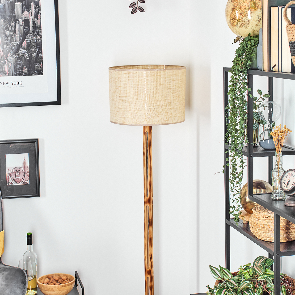 Eine Stehlampe mit beigem Lampenschirm steht neben einem schwarzen Regal mit Pflanzen und Dekoration. Ein gerahmtes New York-Bild hängt an der Wand.