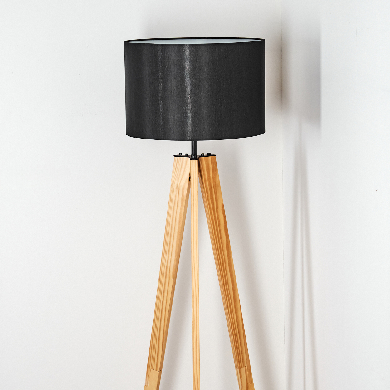 Eine Stehlampe mit schwarzem Lampenschirm und hölzernen Stativbeinen steht vor einer weißen Wand. Die Lampe ist an.