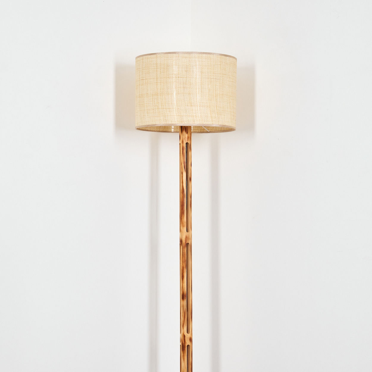 Eine Stehlampe steht vor einer weißen Wand. Sie hat einen beigen Lampenschirm und eine braune Holzstange.