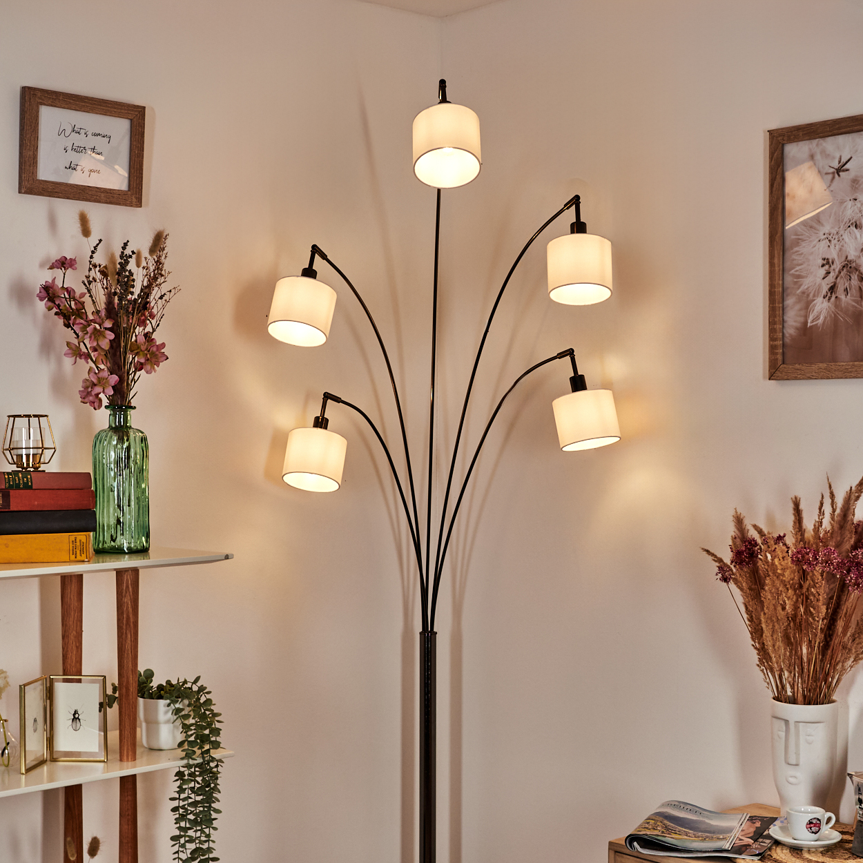 Eine moderne Stehlampe mit sechs weißen Lampenschirmen. Eine Vase mit Blumen und Bildern an der Wand. Ein Tisch mit Büchern.