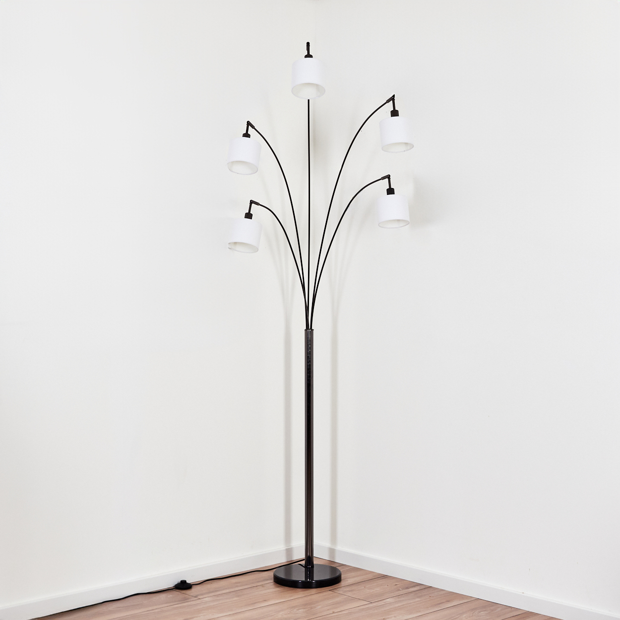 Eine moderne schwarze Stehlampe mit fünf weißen Lampenschirmen. Sie steht auf einem Holzboden vor einer weißen Wand in einer Ecke.