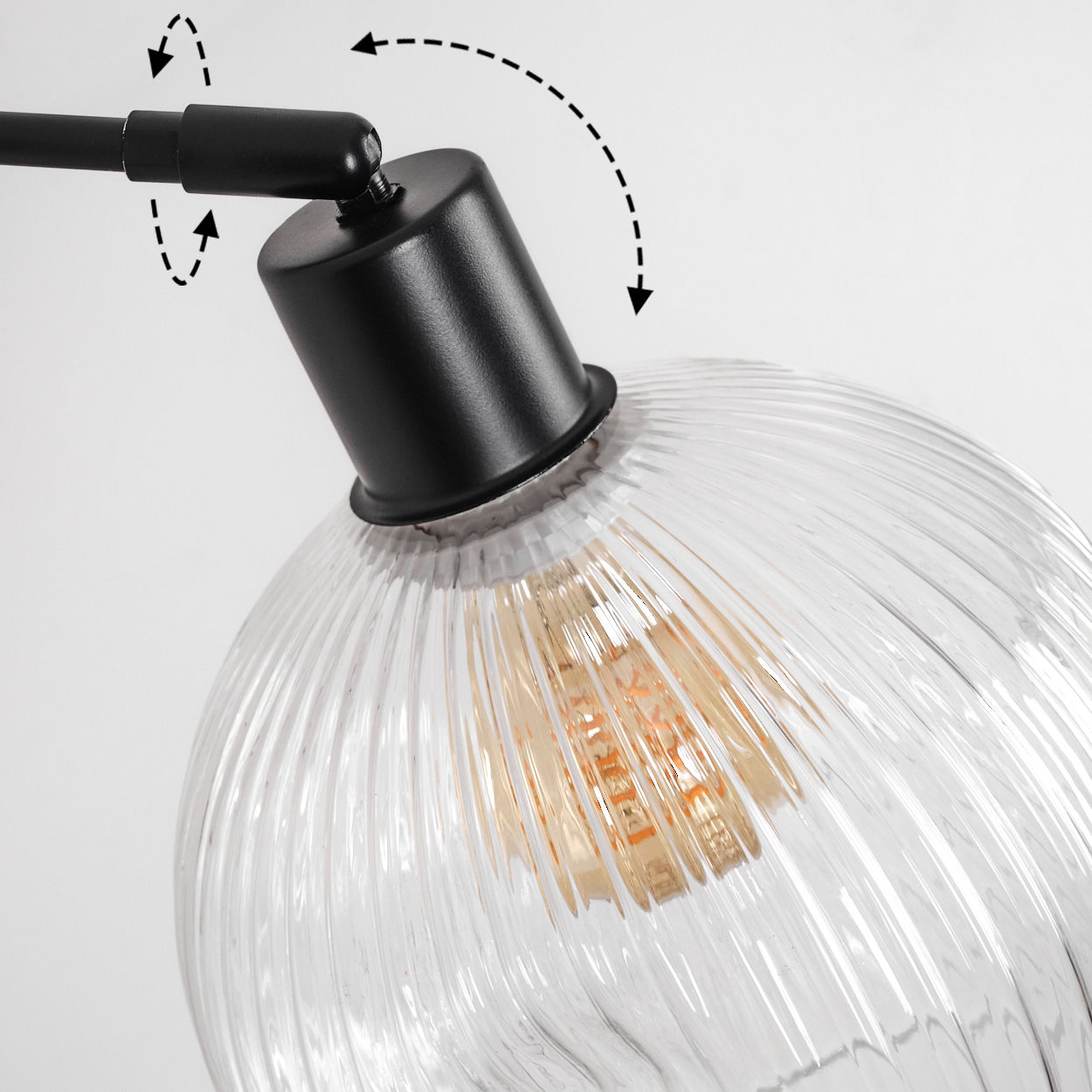 Nahaufnahme einer schwarzen Lampe mit klarem, geripptem Glasschirm. Pfeile zeigen die Verstellbarkeit an. Die Glühbirne ist sichtbar.
