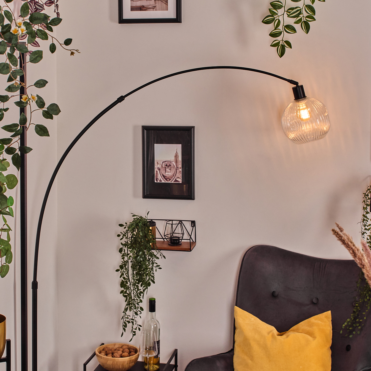 Ein modernes Wohnzimmer mit schwarzer Bogenlampe, grauem Sessel mit gelbem Kissen und dekorativen Pflanzen.