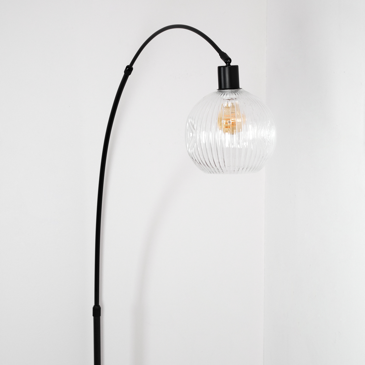 Eine moderne Stehlampe. Sie hat einen schwarzen, gebogenen Arm und einen runden, klaren Glasschirm mit vertikalen Linien. Der Hintergrund ist weiß.