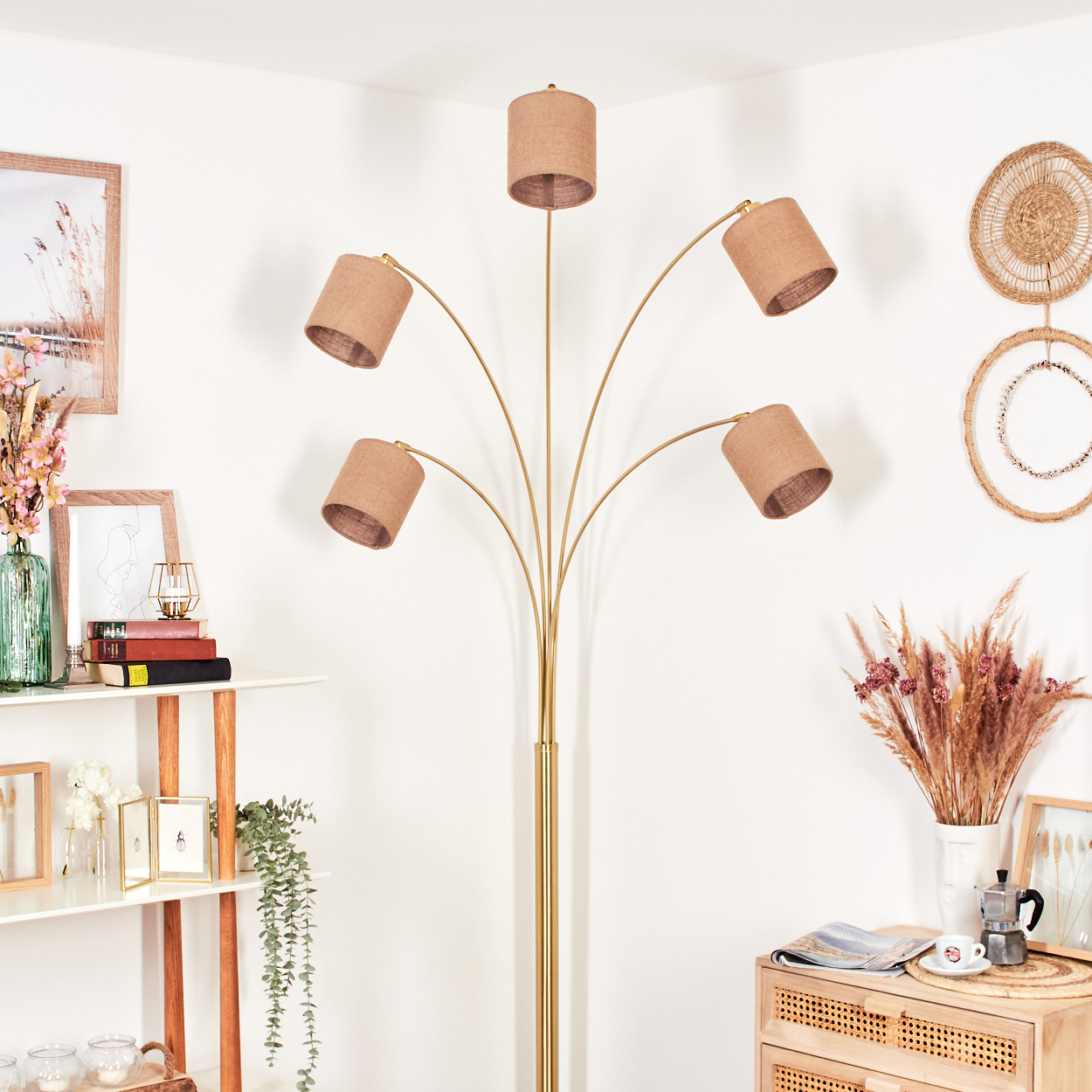 Eine goldene Stehlampe mit fünf Lampenschirmen. Ein Regal mit Büchern, Vasen und Dekorationen. Eine gewebte Wanddekoration. Ein Holzschrank mit Dekor.