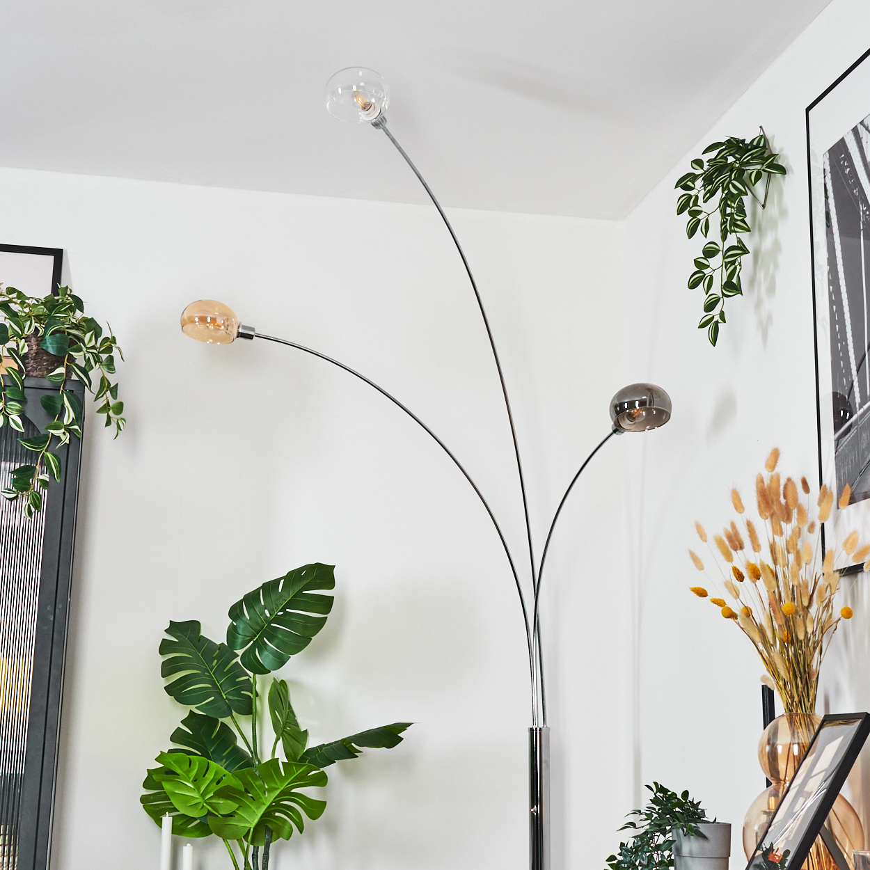 Eine moderne Stehlampe mit drei Glaskugellampen. Pflanzen und gerahmte Kunstwerke umgeben sie. Die Szene spielt sich in Innenräumen mit weißen Wänden ab.