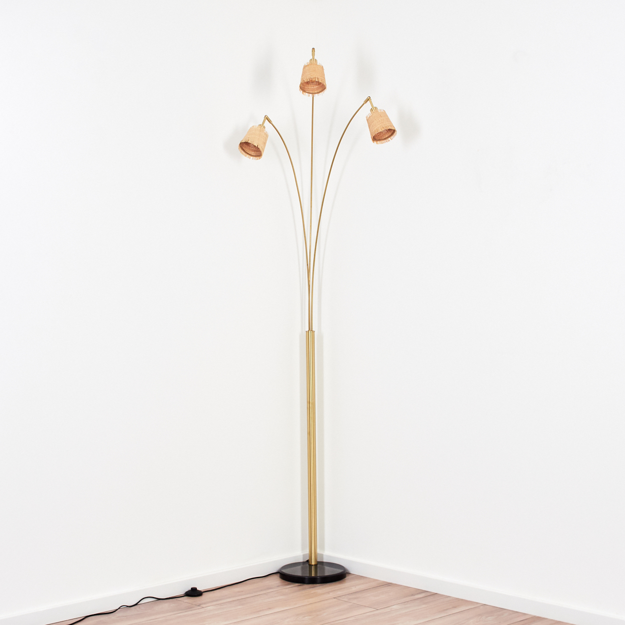 Eine goldene Stehlampe mit drei Lampenschirmen. Sie steht auf einem schwarzen Sockel. Der Hintergrund ist eine weiße Wand und ein Holzboden.