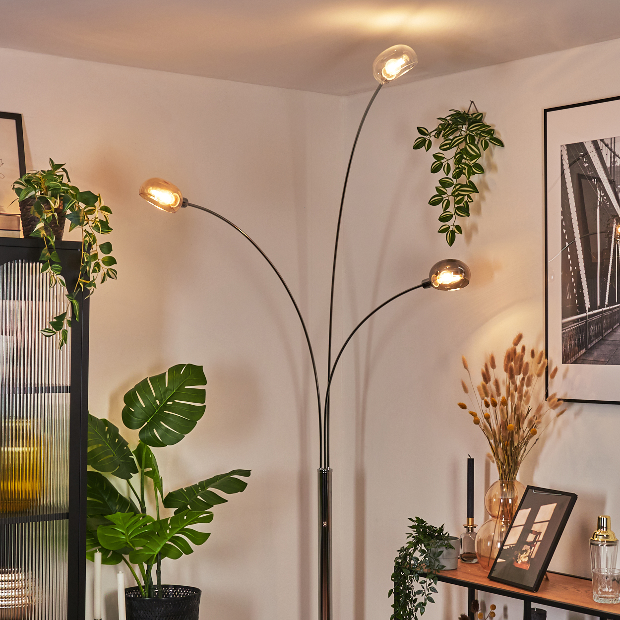 Ein modernes Wohnzimmer mit Stehlampe. Pflanzen und Dekoration sind um die Lampe angeordnet. Ein gerahmtes Bild hängt an der Wand.
