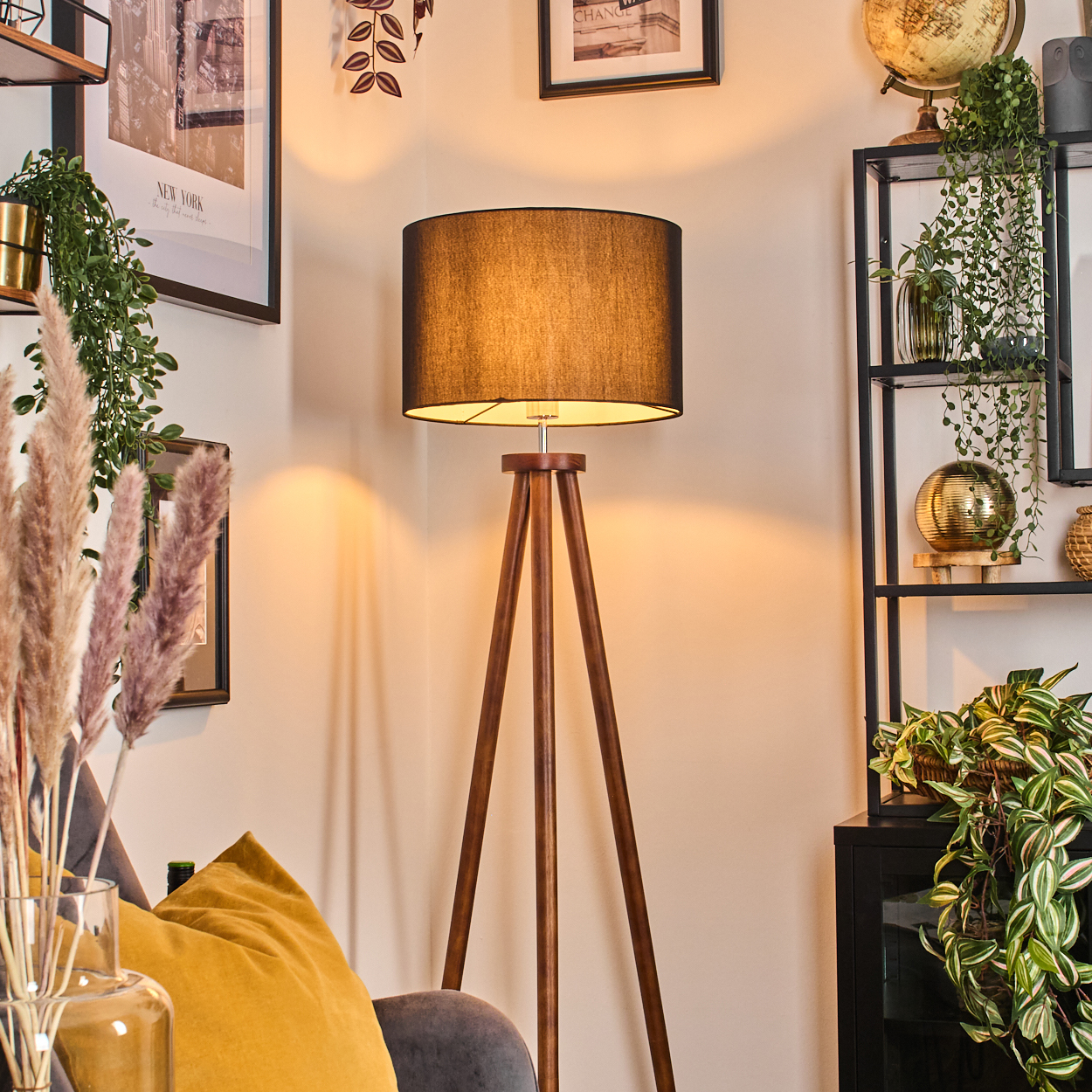 Eine Zimmerecke mit einer Stehlampe, Pflanzen, einer Couch, Bildern und einem Regal mit Deko. Die Lampe ist an.