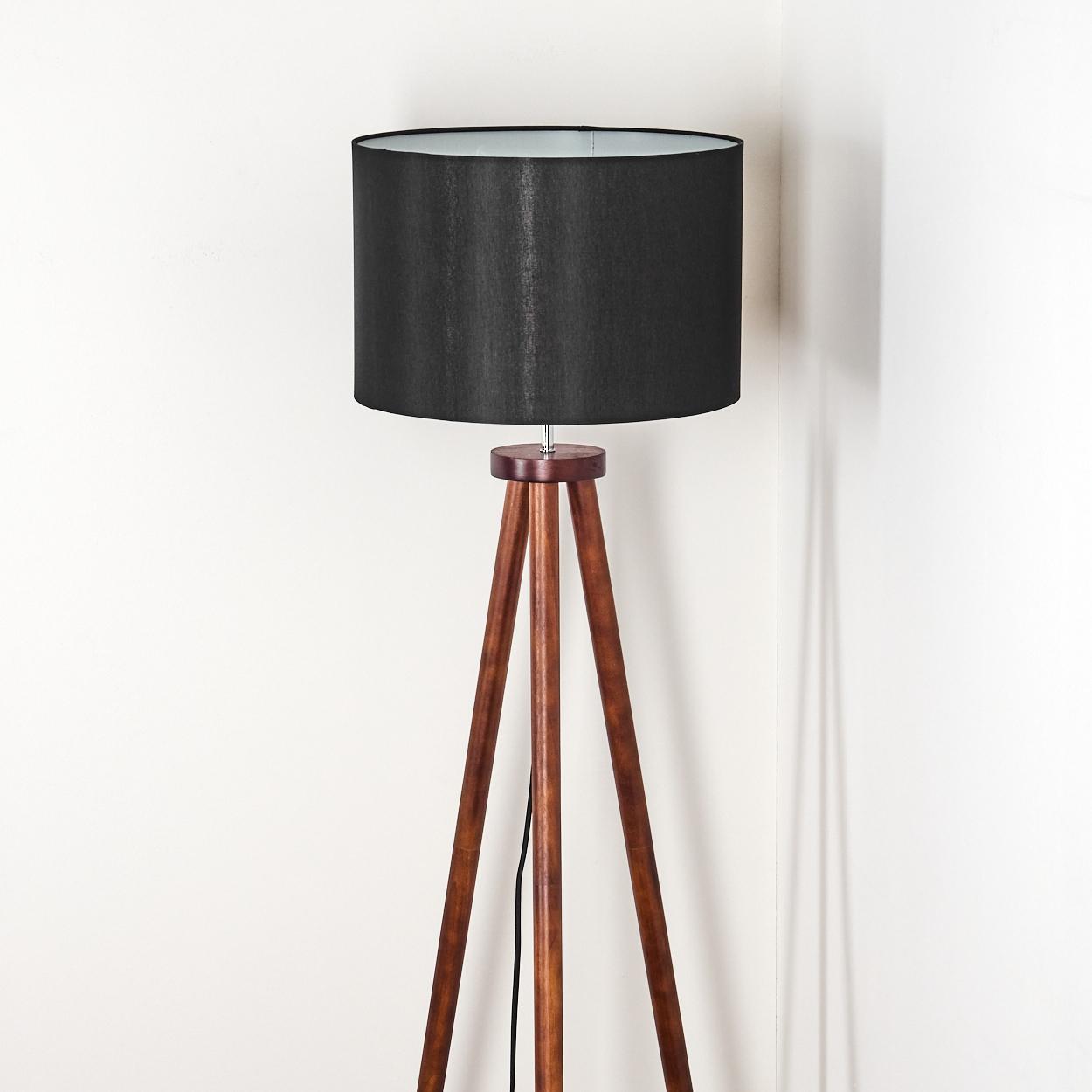 Eine Stehlampe mit Holzstativ und schwarzem Lampenschirm steht vor einer weißen Wand. Die Lampe leuchtet und wirft einen Schatten.