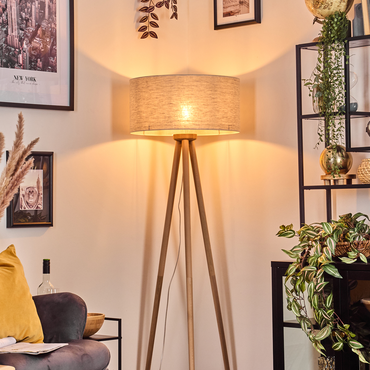 Eine beleuchtete Holzstativlampe steht in einem Raum. Rechts ist ein Regal mit Pflanzen. Bilder und eine Couch sind ebenfalls sichtbar.