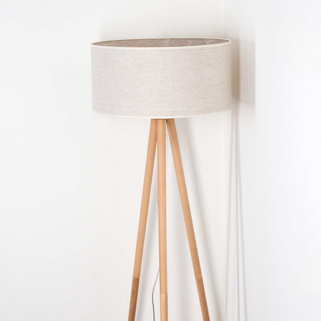 Eine moderne Stehlampe mit beigefarbenem Lampenschirm und Holz-Dreibein-Beinen steht vor einer weißen Wand.