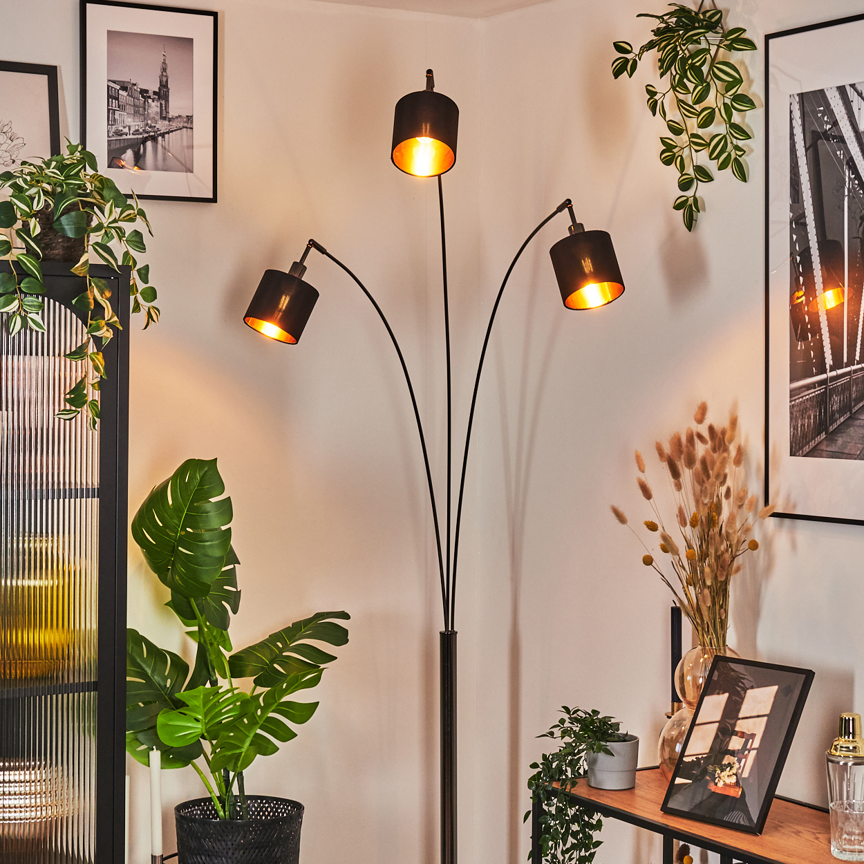 Eine moderne schwarze Stehlampe mit drei Lampenschirmen. Pflanzen und gerahmte Bilder umgeben sie und schaffen eine gemütliche Szene.