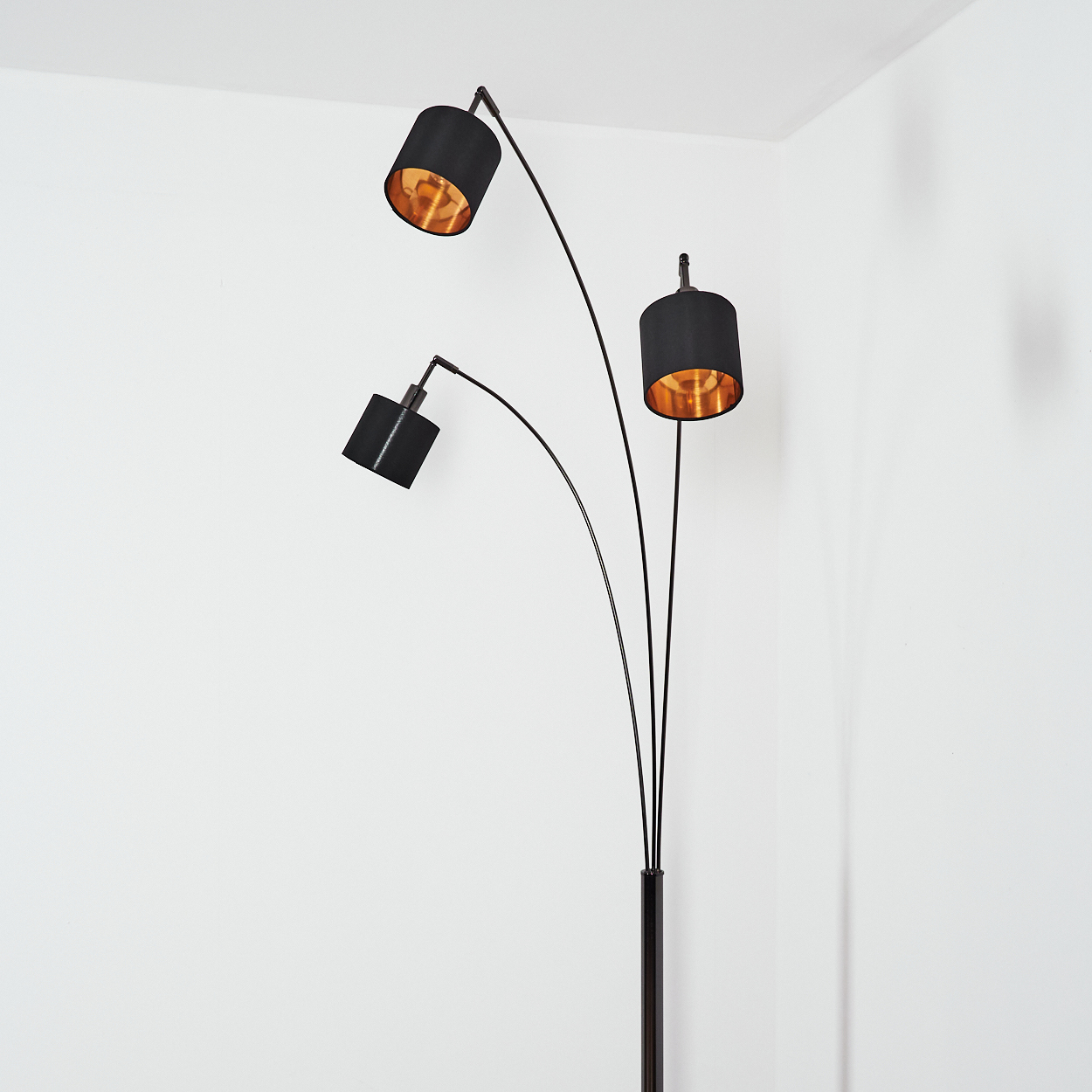 Eine moderne Stehlampe. Sie hat drei schwarze Lampenschirme mit goldenen Innenräumen vor einer weißen Wand in einer Ecke.