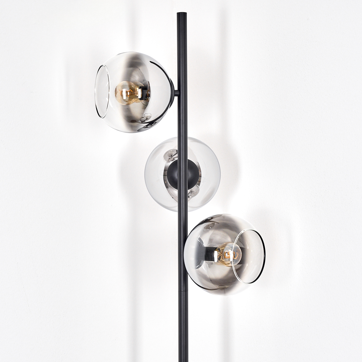 Eine moderne Stehlampe mit drei Glaskugelschirmen. Die Lampe hat einen schwarzen Stab und steht vor einer weißen Wand.