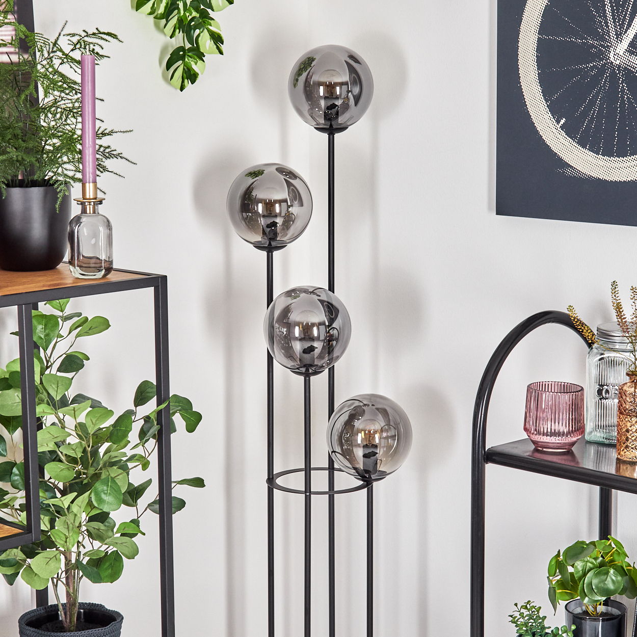 Eine moderne Stehlampe mit vier rauchigen Glaskugeln. Pflanzen und dekorative Regale sind im Hintergrund. Ein Fahrradrad-Kunstwerk ist sichtbar.
