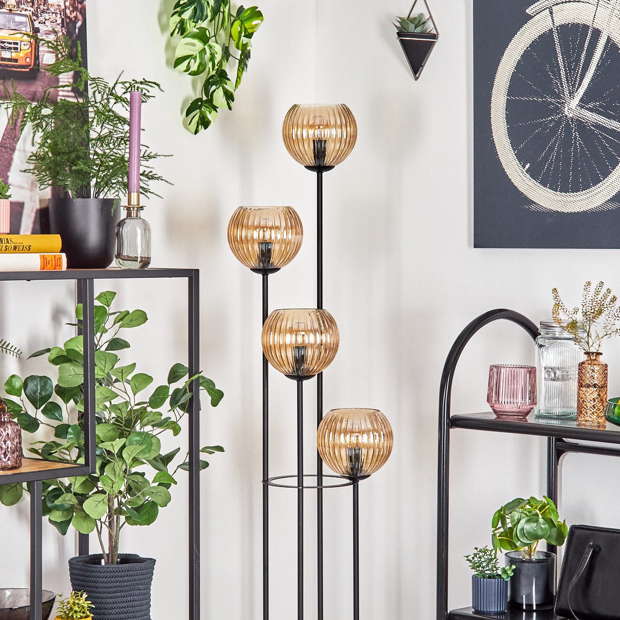 Ein modernes Wohnzimmer mit einem schwarzen Metallregal und einer stilvollen Stehlampe mit bernsteinfarbenen Glasschirmen. Pflanzen und Dekoration sind ebenfalls vorhanden.