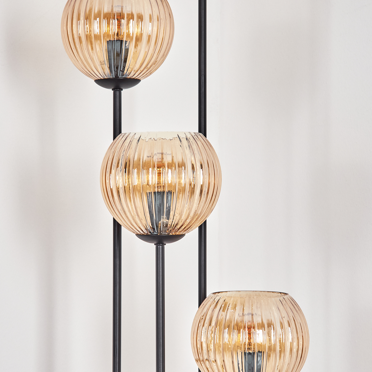 Eine Stehlampe mit drei bernsteinfarbenen, gerippten Glaskugeln. Schwarzes Metall stützt die Kugeln vor einer weißen Wand.