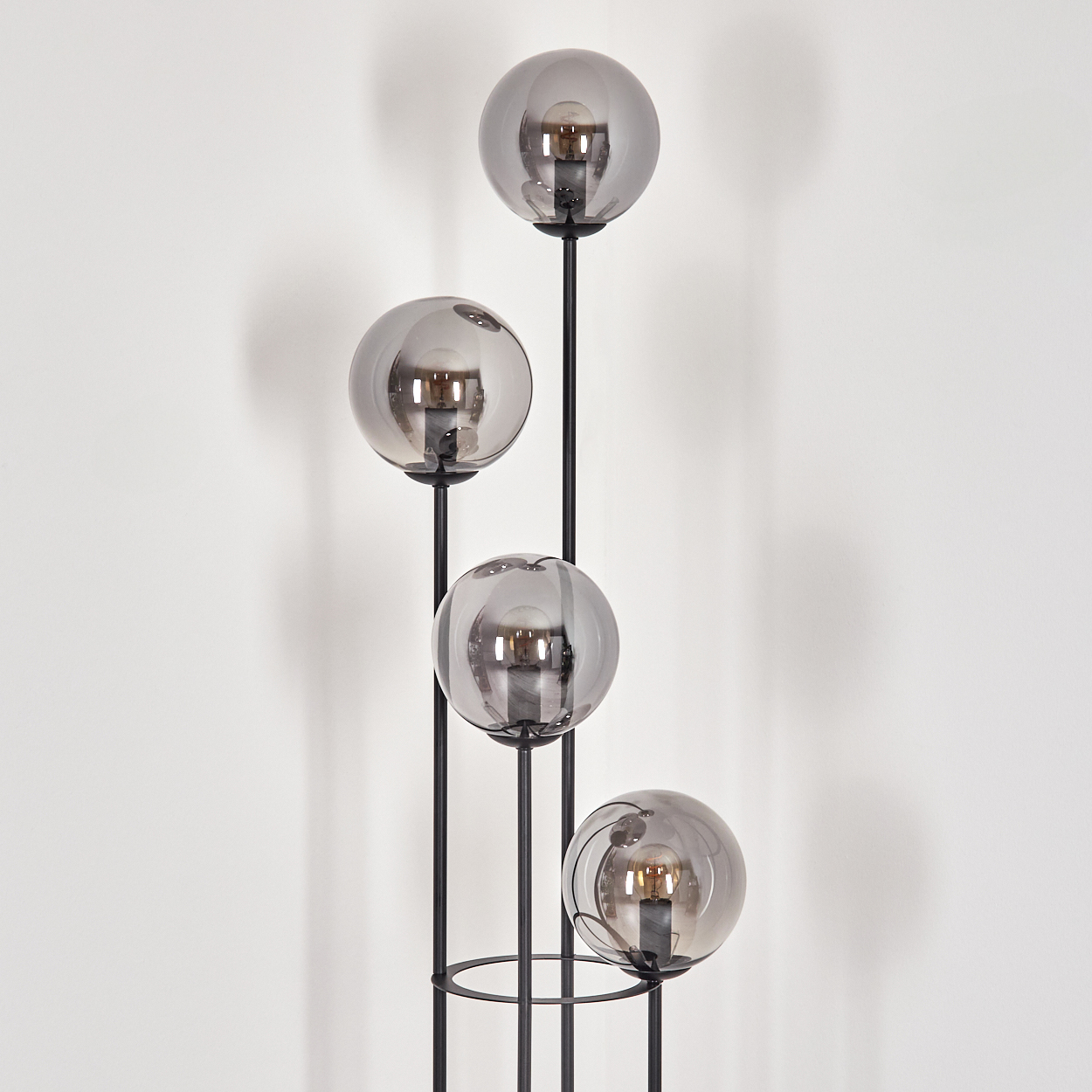 Eine moderne Stehlampe mit vier grauen Glaskugeln auf schwarzen Metallstielen, vor einer weißen Wand.