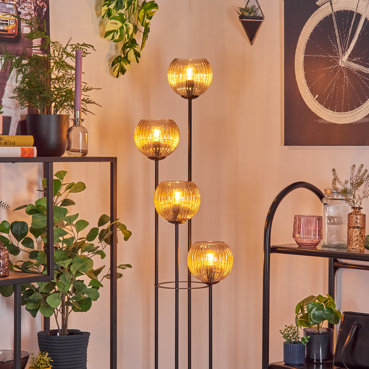 Eine dekorative Lampe mit vier Glaskugeln leuchtet. Pflanzen und Regale mit Dekoration sind im Hintergrund. Die Wand ist hell.