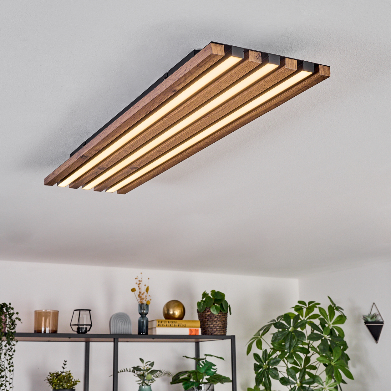 Eine moderne Deckenlampe aus Holz mit integrierten Lichtern. Darunter ein Regal mit Pflanzen und Dekorationsartikeln. Der Hintergrund ist eine weiße Wand.