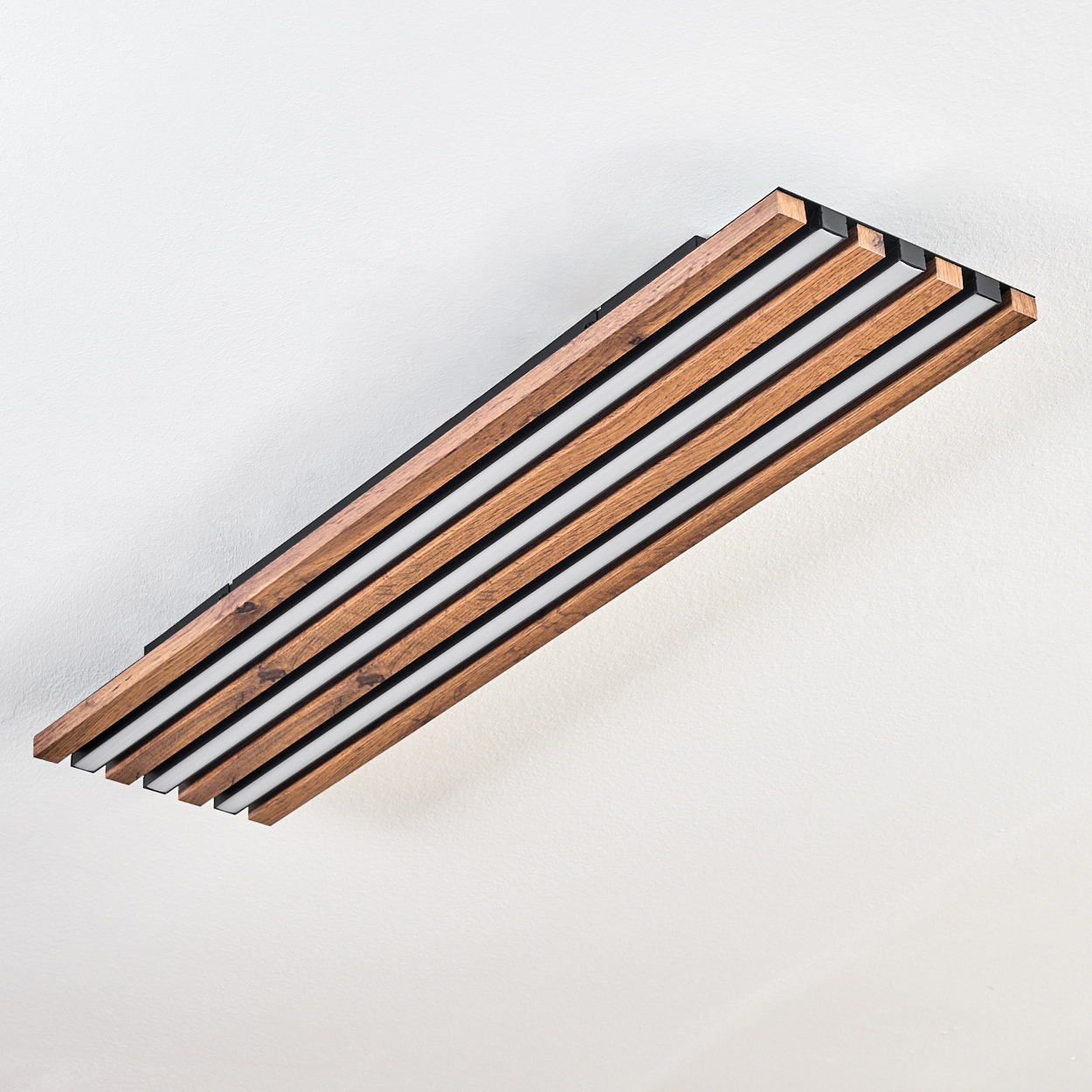 Eine rechteckige Deckenleuchte aus Holz mit schwarzen Kanten und weißen Lichtstreifen. Sie befindet sich an einer weißen Decke.